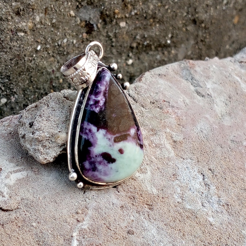May include: A teardrop-shaped pendant with a silver-colored bail and setting. The pendant features a polished stone with a mix of purple, brown, and pale green colors. The stone is set in a decorative silver frame with small silver beads.
