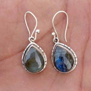 May include: Silver drop earrings featuring teardrop-shaped labradorite stones. The stones show blue, green, and grey tones, set in a detailed silver bezel. The earrings have a hook closure.