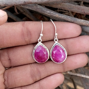 Può includere: Orecchini pendenti in argento con grandi pietre preziose magenta a forma di goccia sfaccettata. Gli orecchini hanno un design semplice con un bordo argentato e chiusura a gancio. Le pietre sono rosa brillante.