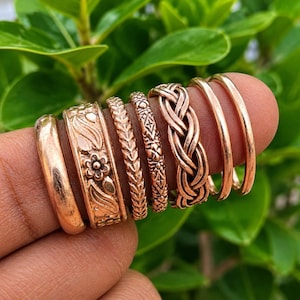 May include: A set of seven copper-toned rings featuring various designs including a smooth band, a floral embossed band, a textured wheat pattern, a geometric etched band, a braided rope design, and two plain thin bands worn on a finger.