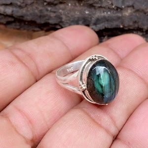 Puede incluir: Un anillo de plata con una gran piedra de labradorita de forma ovalada. La piedra tiene un color verde azulado oscuro con destellos iridiscentes de azul y verde. La banda del anillo está dividida y tiene detalles decorativos. El anillo está marcado con "925".