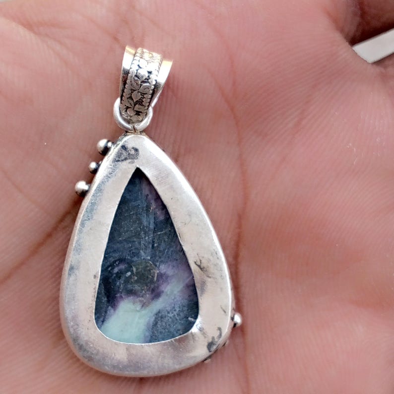 May include: A silver-toned pendant with a teardrop-shaped frame. The pendant features a polished, multi-colored stone with shades of blue, purple, and white. The bail is decorated with floral patterns. Small, round embellishments are placed around the frame.
