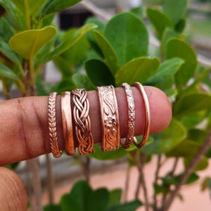 Op de afbeelding: Een set van zes koperkleurige ringen met diverse ontwerpen, waaronder een visgraatpatroon, een gladde band, een gevlochten textuur, een band met bloemreliëf, een klein geometrisch patroon en een eenvoudige dunne band, op een rij.