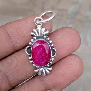 May include: A silver pendant featuring an oval, deep red gemstone. The gemstone is set in a decorative silver frame with a scalloped top and bottom design. The pendant has a small loop at the top for a chain.