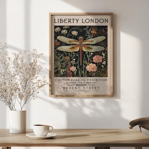 May include: Framed vintage advertisement for Liberty London's Cotton Fabrics Exhibition. The artwork features a dragonfly with detailed wings, surrounded by flowers and foliage. The poster includes text about the exhibition and location.