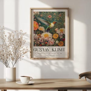 Op de afbeelding: Een ingelijste kunstdruk met een kleurrijke kolibrie in vlucht te midden van een bloementuin. De print is getiteld "GUSTAV KLIMT" met extra tekst eronder. Het kunstwerk wordt aan een muur getoond.