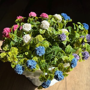 Pode incluir: Um arranjo floral decorativo num vaso de cerâmica branco. O arranjo apresenta uma variedade de flores coloridas, incluindo flores rosa, brancas, azuis e roxas, num fundo de folhas verdes. O vaso está sobre uma superfície de madeira.