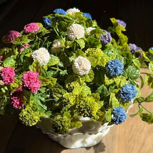 May include: A decorative floral arrangement in a white ceramic bowl. The arrangement features a variety of colorful artificial flowers, including pink, blue, white, and purple blooms, set against green foliage. The bowl has a scalloped edge.