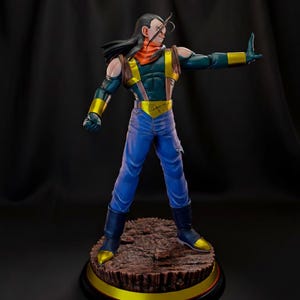 May include: A detailed figurine of Android 17 from Dragon Ball Z. The figure is posed with one arm raised, wearing a blue and gold outfit, with a red scarf and long black hair. The figure stands on a brown base with a gold trim.