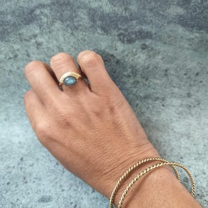 Peut inclure: Une bague en or ornée d'une pierre ovale aux reflets bleu-gris irisés montée sur un anneau doré en demi-cercle, accompagnée de deux bracelets fins en fil d'or torsadé portés au poignet, présentant une finition métallique et texturée.