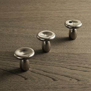 May include: Three polished silver cabinet knobs, each with a mushroom-shaped design. The knobs have a rounded top and a cylindrical base. They are displayed on a wooden surface with a visible grain.