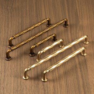 May include: A collection of brass cabinet pulls with a textured, diamond-patterned design. The pulls are arranged on a wooden surface, showcasing their varying sizes and warm, metallic finish. These decorative hardware pieces are ideal for kitchen or bathroom cabinets.
