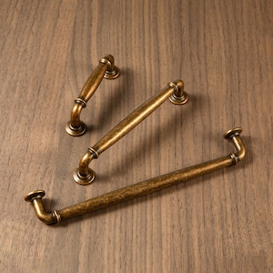 May include: Three antique brass cabinet pulls in varying sizes are arranged on a wooden surface. The pulls have a curved design with rounded ends and a distressed finish, suggesting a vintage aesthetic. The hardware is ideal for kitchen or bathroom cabinets.