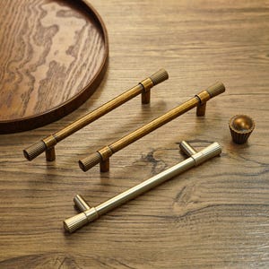 May include: A selection of bronze and brass cabinet hardware, including two long bar pulls, a shorter pull, and a small knob. The hardware has a ribbed design and is displayed on a wooden surface, with a wooden tray.