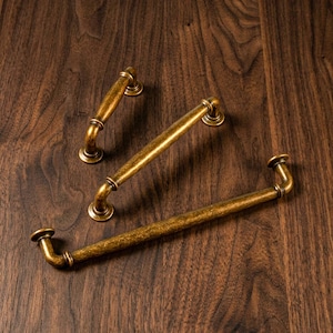 Puede incluir: Tres tiradores de armario de latón antiguo con acabado envejecido dispuestos sobre una superficie de madera oscura. Los tiradores tienen un diseño curvo con extremos redondeados y una barra central. El herraje es ideal para añadir un toque vintage.