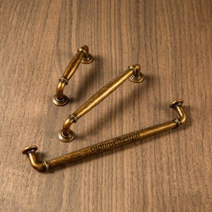 Può includere: Tre maniglie per mobili in ottone antico su una superficie di legno. Le maniglie sono di diverse dimensioni e hanno un design curvo. Il testo "OldtimeDesain" è visibile sulla maniglia più grande.