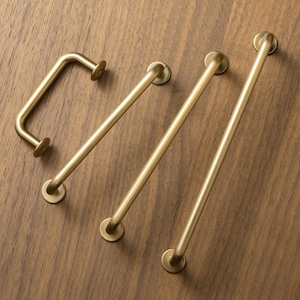 May include: Four brass cabinet pulls in different lengths and shapes. The hardware includes a curved handle and three straight bar pulls, all with circular mounting plates. The pulls are arranged on a wooden surface.