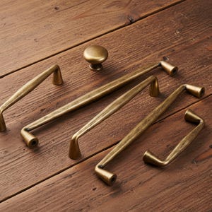 Op de afbeelding: Een verzameling antieke messing kastbeslagen, waaronder verschillende handgrepen en een knop. De handgrepen variëren in grootte en vorm, met een gebogen ontwerp. De hardware is geplaatst op een rustiek houten oppervlak.