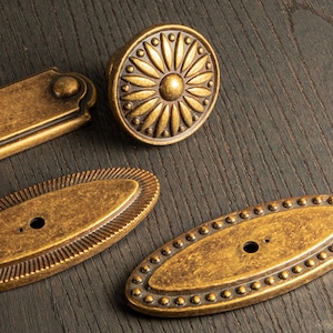 Peut inclure: Gros plan sur des pi&egrave;ces de quincaillerie en laiton antique, dont un bouton rond avec un motif floral et plusieurs tirettes ovales et rectangulaires. La quincaillerie a une finition vieillie, sugg&eacute;rant un aspect vieilli. Les pi&egrave;ces sont dispos&eacute;es sur une surface en bois fonc&eacute;.