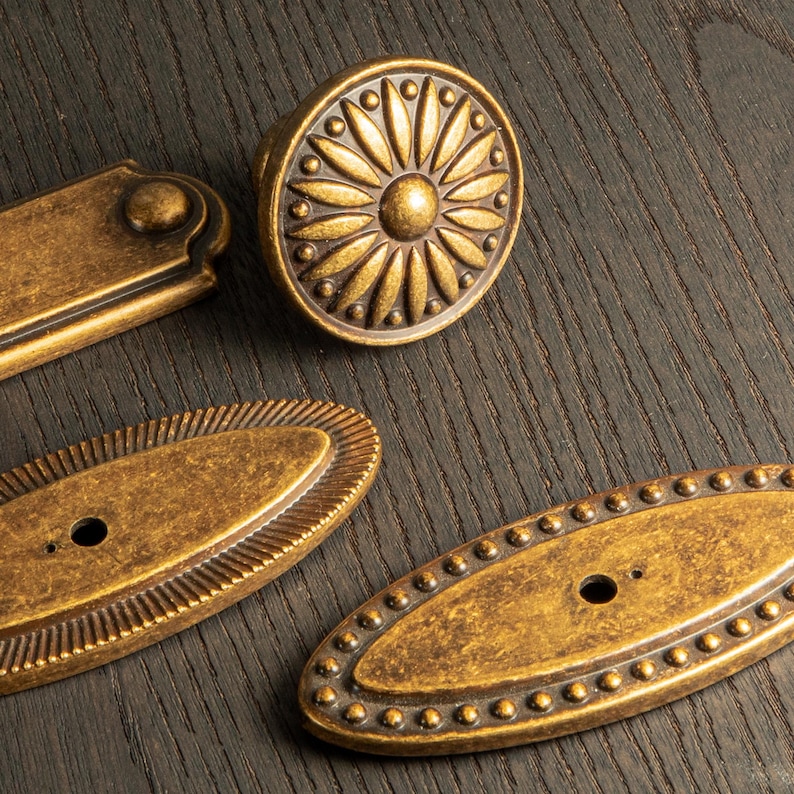 Peut inclure: Gros plan sur des pi&egrave;ces de quincaillerie en laiton antique, dont un bouton rond avec un motif floral et plusieurs tirettes ovales et rectangulaires. La quincaillerie a une finition vieillie, sugg&eacute;rant un aspect vieilli. Les pi&egrave;ces sont dispos&eacute;es sur une surface en bois fonc&eacute;.
