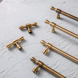 May include: A selection of antique brass cabinet handles and knobs. The knobs feature a T-bar design, and the handles are cylindrical with decorative ends. The hardware is arranged on a textured, light grey surface.