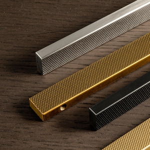 Può includere: Primo piano di diverse maniglie per mobili in metallo argentato, dorato e nero. Ogni maniglia ha una superficie strutturata a forma di diamante e una forma rettangolare. Le maniglie sono disposte diagonalmente su una superficie di legno scuro.