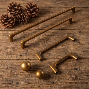 Può includere: Una collezione di ferramenta per mobili in ottone antico, tra cui diverse maniglie e due pomelli sferici. Le maniglie variano di dimensioni e presentano un design curvo. L'immagine include anche pigne, il tutto su una superficie di legno rustico.
