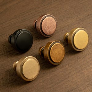 Może przedstawiać: Pięć okrągłych gałek do szafek w różnych metalicznych wykończeniach: czarnym, miedzianym, mosiężnym i antycznym mosiądzu. Każda gałka ma okrągłą powierzchnię i krótki trzpień do montażu. Gałki są ułożone na drewnianej powierzchni.