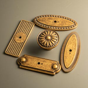 Peut inclure: Un assortiment de quincaillerie de meubles en laiton antique, comprenant des boutons et des poign&eacute;es. La quincaillerie pr&eacute;sente diverses formes, telles qu'ovales, rectangulaires et rondes, avec des d&eacute;tails d&eacute;coratifs tels que des bords perl&eacute;s et des motifs floraux.
