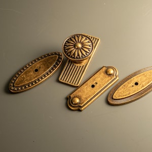 Peut inclure: Divers &eacute;l&eacute;ments de quincaillerie en laiton antique, dont deux plaques ovales avec des bords perl&eacute;s, une plaque rectangulaire et un bouton de meuble avec un motif floral. L'ensemble est dispos&eacute; sur une surface vert p&acirc;le.