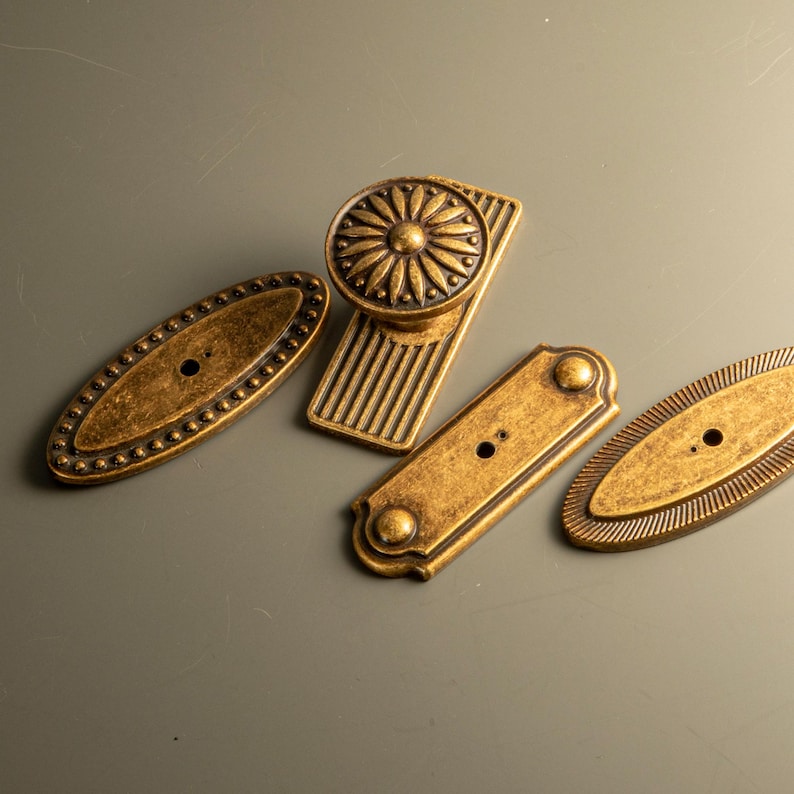 Peut inclure: Divers &eacute;l&eacute;ments de quincaillerie en laiton antique, dont deux plaques ovales avec des bords perl&eacute;s, une plaque rectangulaire et un bouton de meuble avec un motif floral. L'ensemble est dispos&eacute; sur une surface vert p&acirc;le.