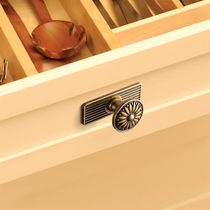 Peut inclure: Gros plan sur un tiroir de cuisine avec une poign&eacute;e d&eacute;corative en laiton. La poign&eacute;e a une plaque arri&egrave;re rectangulaire avec des lignes horizontales et un bouton rond &agrave; motifs floraux. Une cuill&egrave;re en bois est visible dans le tiroir ouvert.