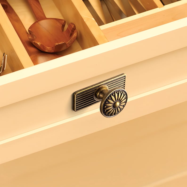 Peut inclure: Gros plan sur un tiroir de cuisine avec une poign&eacute;e d&eacute;corative en laiton. La poign&eacute;e a une plaque arri&egrave;re rectangulaire avec des lignes horizontales et un bouton rond &agrave; motifs floraux. Une cuill&egrave;re en bois est visible dans le tiroir ouvert.