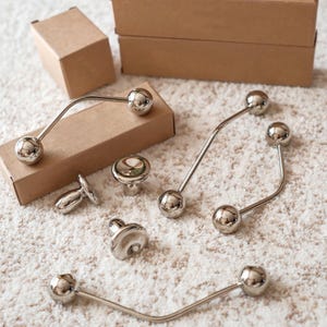 Peut inclure: Une collection de quincaillerie d'armoire argentée brillante, comprenant des boutons et des poignées. Le matériel a un design moderne avec des extrémités sphériques et des barres courbes ou droites. Les articles sont disposés sur une surface blanche texturée, avec des boîtes en carton en arrière-plan.