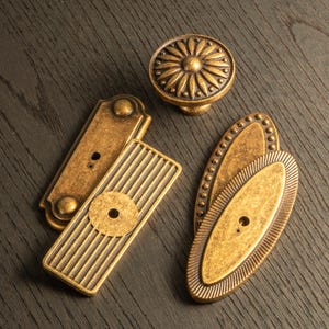 Puede incluir: Un surtido de herrajes de latón antiguo para armarios, que incluye un pomo redondo con diseño floral, dos tiradores rectangulares y dos tiradores ovalados con bordes decorativos. Los herrajes están dispuestos sobre una superficie de madera oscura.