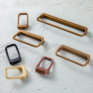 Può includere: Un assortimento di maniglie rettangolari per mobili in varie finiture metalliche, tra cui bronzo, oro e nero. L'hardware è esposto su una superficie chiara, mostrando le diverse forme e colori.