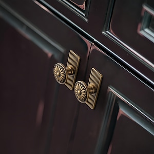 Peut inclure: Gros plan sur deux boutons de meuble en laiton antique avec un motif de soleil. Chaque bouton est mont&eacute; sur une plaque arri&egrave;re rectangulaire avec des lignes verticales. Les boutons sont fix&eacute;s sur une porte d'armoire sombre et brillante.