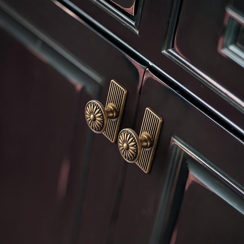 Peut inclure: Gros plan sur deux boutons de meuble en laiton antique avec un motif de soleil. Chaque bouton est mont&eacute; sur une plaque arri&egrave;re rectangulaire avec des lignes verticales. Les boutons sont fix&eacute;s sur une porte d'armoire sombre et brillante.