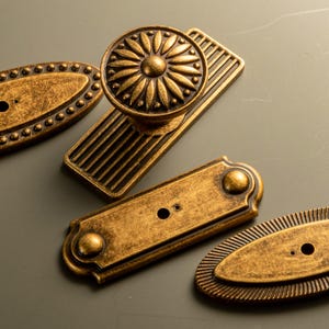 Peut inclure: Gros plan sur une quincaillerie de porte en laiton antique. L'ensemble comprend un bouton de porte rond avec un motif floral, une plaque arri&egrave;re rectangulaire et deux plaques ovales et rectangulaires avec des d&eacute;tails d&eacute;coratifs.