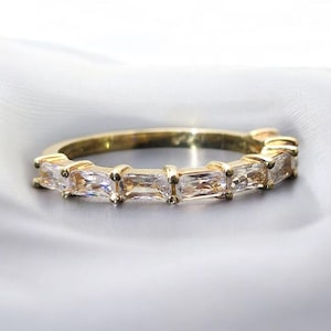 May include: A gold ring with a line of rectangular, clear gemstones. The ring's band is gold, and the gemstones are set closely together, creating a continuous sparkle. The ring is displayed on a soft, white surface.