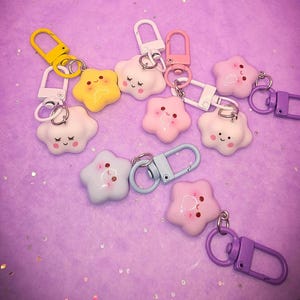 Puede incluir: Una colección de llaveros en forma de estrella en varios colores pastel, incluyendo amarillo, rosa y azul. Cada llavero presenta una cara sonriente y un cierre metálico. Los llaveros están dispersos sobre una superficie púrpura.