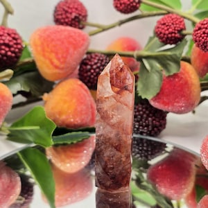May include: A clear quartz crystal point with reddish-brown inclusions, reflecting light. The crystal is displayed on a mirrored surface, surrounded by artificial peaches, berries, and green leaves. The crystal is a natural, earthy item.