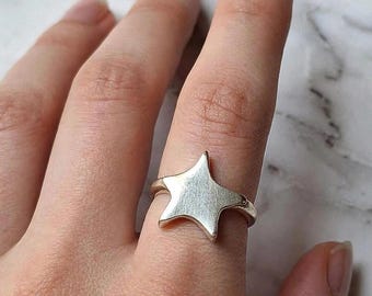 Puede incluir: Un anillo de tono plateado con un diseño de estrella de cinco puntas. La estrella tiene un acabado liso y mate, centrado en la banda que rodea el dedo. La superficie metálica refleja la luz de manera uniforme sobre la forma geométrica.