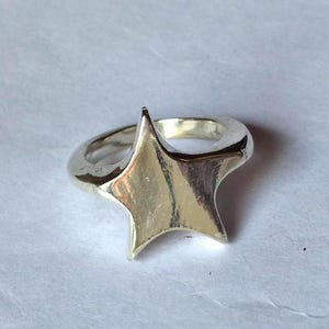 Op de afbeelding: Een zilverkleurige ring met een stervormig ontwerp. De ster heeft vijf punten en is het middelpunt van de ring. De ringband is glad en afgerond, en de ster is verhoogd.