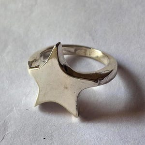 Op de afbeelding: Een zilverkleurige ring met een vijfpunts sterontwerp. De ster is het middelpunt, met een glad, gepolijst oppervlak. De ringband is ook zilverkleurig, wat een samenhangende uitstraling cre&euml;ert.