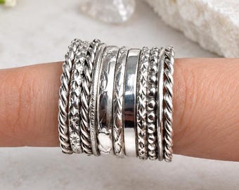 May include: A set of ten silver-toned stacking rings worn on a finger. The collection includes various textures such as twisted rope patterns, beaded bands, smooth polished surfaces, and intricate engraved designs, all crafted from a metallic material.