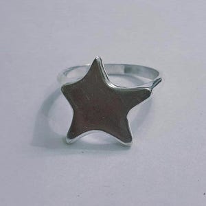 Op de afbeelding: Een zilverkleurige ring met een stervormig ontwerp. De ster is het middelpunt, met een glad, metalen oppervlak. De ringband is eenvoudig en past bij de vorm van de ster. Dit sieraad is geschikt voor dagelijks gebruik.