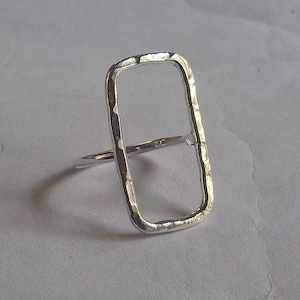 May include: A silver ring with a rectangular open design. The ring has a hammered texture on the frame, giving it a rustic look. The band is thin and smooth, contrasting with the textured rectangle. The ring is set against a neutral background.