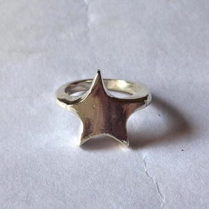 Op de afbeelding: Een zilverkleurige ring met een sterontwerp. De ster, met vijf punten, is het centrale element. De ring staat tegen een neutrale achtergrond, wat de metaalglans en de vorm van de ster benadrukt.
