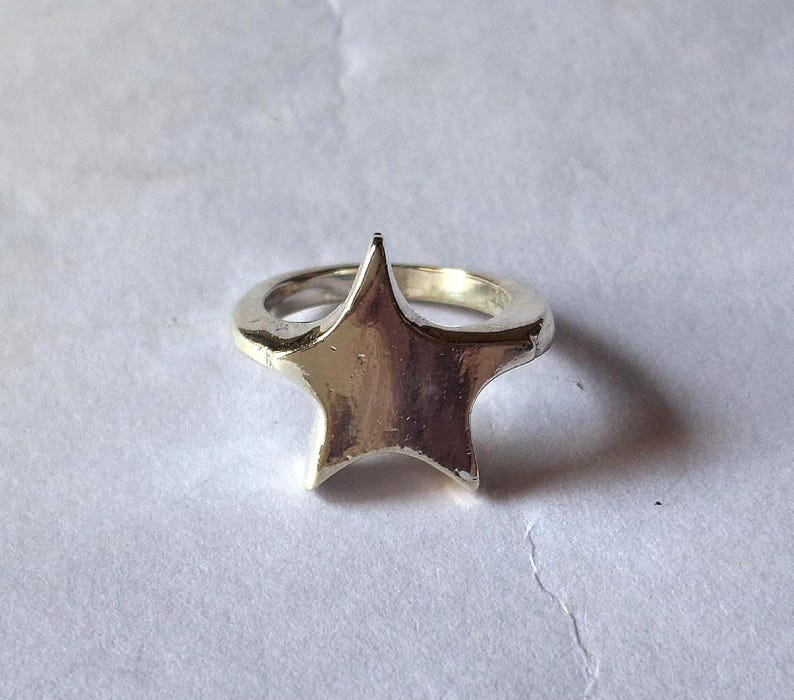 Op de afbeelding: Een zilverkleurige ring met een sterontwerp. De ster, met vijf punten, is het centrale element. De ring staat tegen een neutrale achtergrond, wat de metaalglans en de vorm van de ster benadrukt.